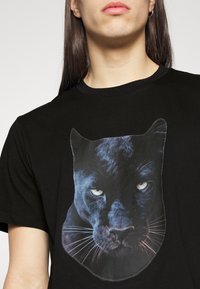 Persona con una camiseta negra con un gran y detallado gráfico de la cara de un pantera negra centrado en el pecho, sobre un fondo liso.