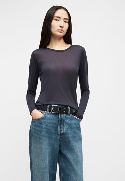 Sieviete, tērpusies caurspīdīgā tumšā garroku krekliņā, kas ielikts vaļīgās zilās džinsu biksēs ar melnu jostu, stāv ar rokām kabatās.