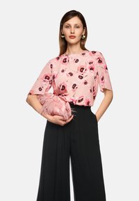 Blumenbluse in rosa Tönen mit kurzen Ärmeln, kombiniert mit weiten schwarzen Hosen. Hält eine rosa Feder-Clutch.