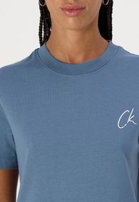 Camiseta de algodón de color azul claro con cuello redondo y mangas cortas, que cuenta con un logotipo bordado en blanco "CK" en la parte izquierda del pecho.