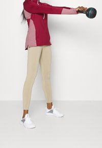 Bordeaux sporttopp med rosa detaljer, långärmad och med sidslitsar. Beige leggings och vita sneakers. Personen håller en kettlebell.