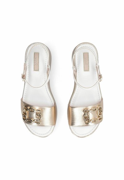 Sandales blanches avec des brides métalliques dorées ornées de chaînes en or, vues de dessus sur un fond blanc.