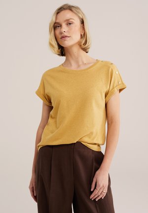 Blondynka w musztardowo-żółtej bluzce z krótkim rękawem i guzikami oraz ciemnobrązowych spodniach z wysokim stanem na neutralnym tle.