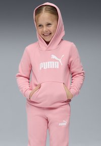 Rožinė maikutė su gobtuvu ir atitinkančios sportinės kelnės, pagamintos iš švelnaus audinio, su dideliu baltu Puma logotipu priekyje ir šone. Maikutė turi kengūrišką kišenę.
