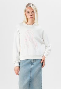 Pullover bianco con collo a giro e vestibilità oversize, caratterizzato da una grafica rosa. Indossato con una gonna di denim blu chiaro.