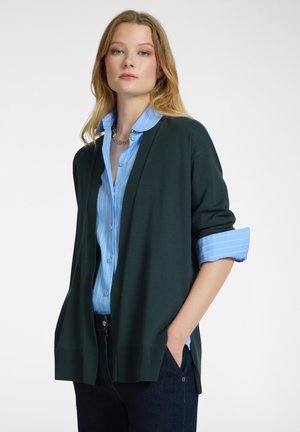 Una giovane donna con capelli biondi indossa un cardigan aperto verde scuro su una camicia a righe blu chiaro con maniche arrotolate e jeans scuri.