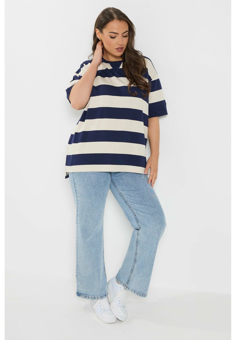 Femme aux longs cheveux bruns portant une chemise oversize rayée bleu marine et blanc, un jean bleu clair et des baskets blanches, debout contre un mur blanc.