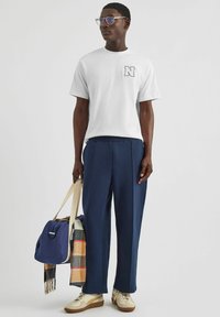 Biała bawełniana koszulka z logo, granatowe spodnie z zakładkami, beżowe trampki, przezroczyste okulary, trzymając niebieską torbę na ramię i szkocki szalik.