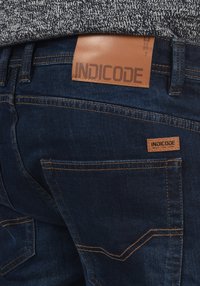 Tamno plave traperice izrađene od denima, s smešnim kožnim flasterom na kojem piše "INDICODE" i malom fabric tagom u blizini džepa.