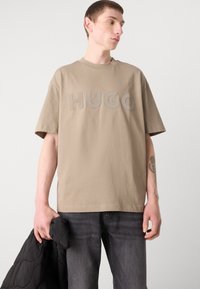Krekls ar īsām piedurknēm brūnā krāsā ar lieliem "HUGO" burtiem, kas ir saskaņotā krāsā. Modelis tur melnu nošuvumu jaku. Izbalējušas pelēkas džinsas papildina apģērbu.