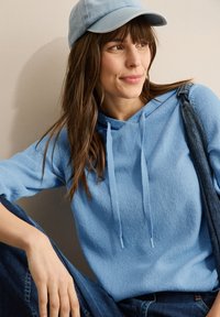 Felpa con cappuccio azzurro chiaro con lacci, tessuto strutturato, indossata con salopette in denim, abbinata a un cappellino azzurro chiaro.