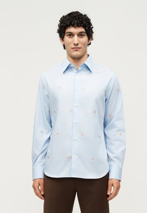 Άνδρας με σγουρά μαλλιά που φοράει ανοιχτό μπλε πουκάμισο με μακριά μανίκια και μικρά λουλουδάτα σχέδια, συνδυασμένο με σκούρο καφέ παντελόνι.