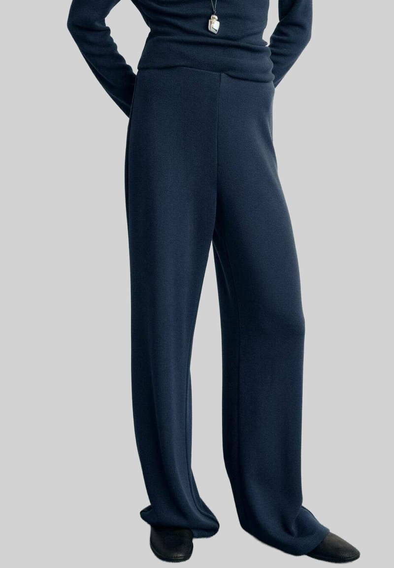 Personne portant un pantalon large bleu marine foncé, des chaussures noires et un haut à manches longues assorti bleu marine foncé avec un collier pendentif.