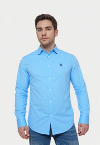 Camisa de botones de manga larga en azul claro, con un diseño ajustado y un pequeño logo bordado en el pecho, combinada con vaqueros azules.