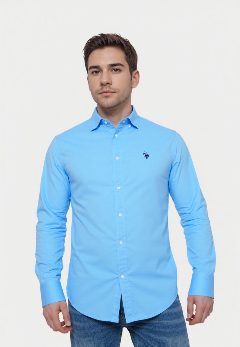 Camisa de botones de manga larga en azul claro, con un diseño ajustado y un pequeño logo bordado en el pecho, combinada con vaqueros azules.
