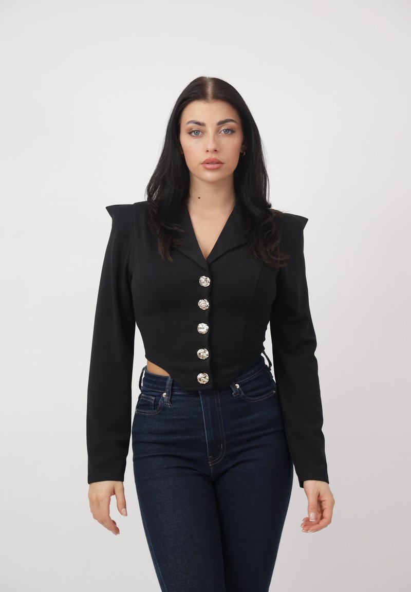 Frau mit langen, dunklen Haaren, die eine schwarze, kurze Jacke mit großen silbernen Knöpfen und dunkle hoch taillierte Jeans trägt, steht vor einem weißen Hintergrund.