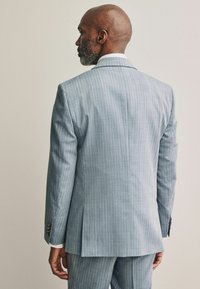 Costume bleu clair à rayures avec un tissu texturé. Les caractéristiques notables incluent une fente unique, des poignets boutonnés et une chemise habillée blanche en dessous.