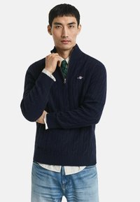 Marineblauer Strickpullover mit einem Viertelreißverschluss, der ein Kabelmuster und gerippte Bündchen und Saum aufweist, getragen über einem karierten Hemd und einer Krawatte.