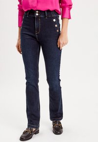 Dark blue flared jeans with a high waist, featuring three silver buttons on the left side, and a smooth denim texture. Paired with black loafers.