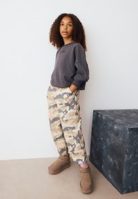 Sweat-shirt gris à coupe ample avec poignets côtelés, associé à un pantalon camouflage beige et gris. Bottines marron avec des accents roses à proximité.