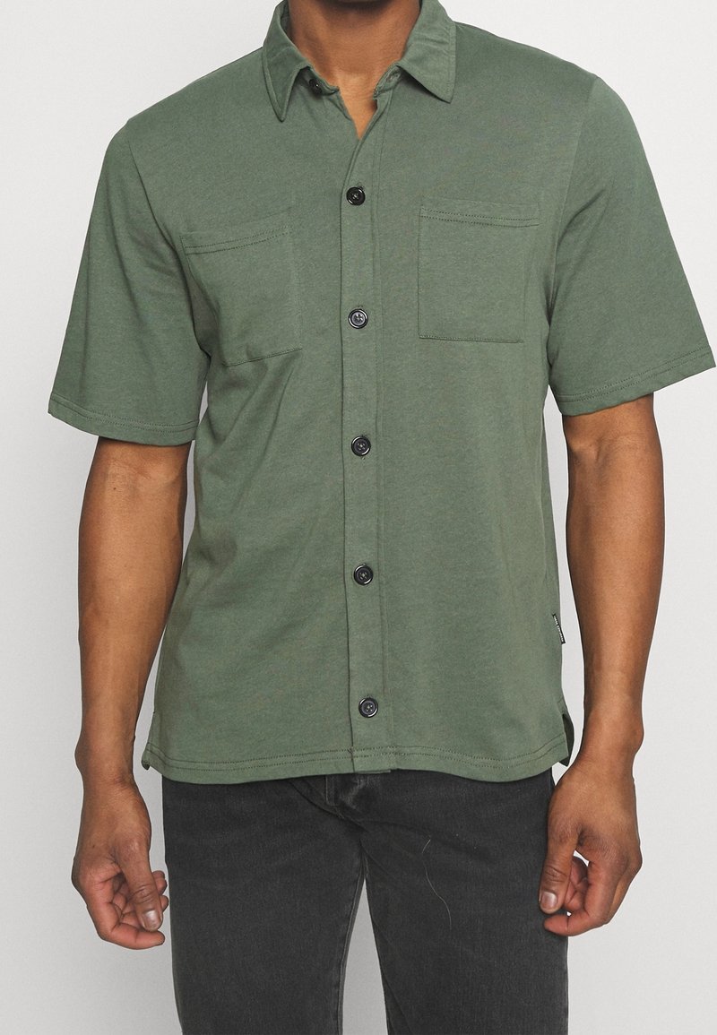 Chemise verte à manches courtes en tissu doux, avec deux poches poitrine et boutons noirs sur le devant pour la fermeture.
