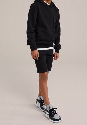 Dreng iført en sort hættetrøje, sorte shorts, grå sokker og sort-hvide sneakers, der står foran en ensfarvet baggrund.