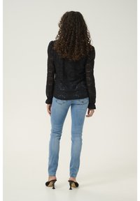 Top in pizzo nero con maniche lunghe e spalle a sbuffo, abbinato a jeans aderenti blu chiaro e scarpe con tacco nere. Capelli ricci visibili.