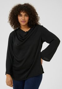 Sort langærmet top med draperet halsudskæring og tekstureret stof. Har let udvidede ærmer og en afslappet pasform, parret med blå jeans.
