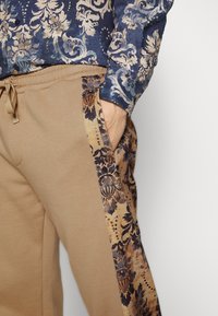 Beiga sweatpants har en mönstrad sidopanel i nyanser av brunt och blått, tillsammans med en blå blommönstrad skjorta. Finns med dragsko i midjan.