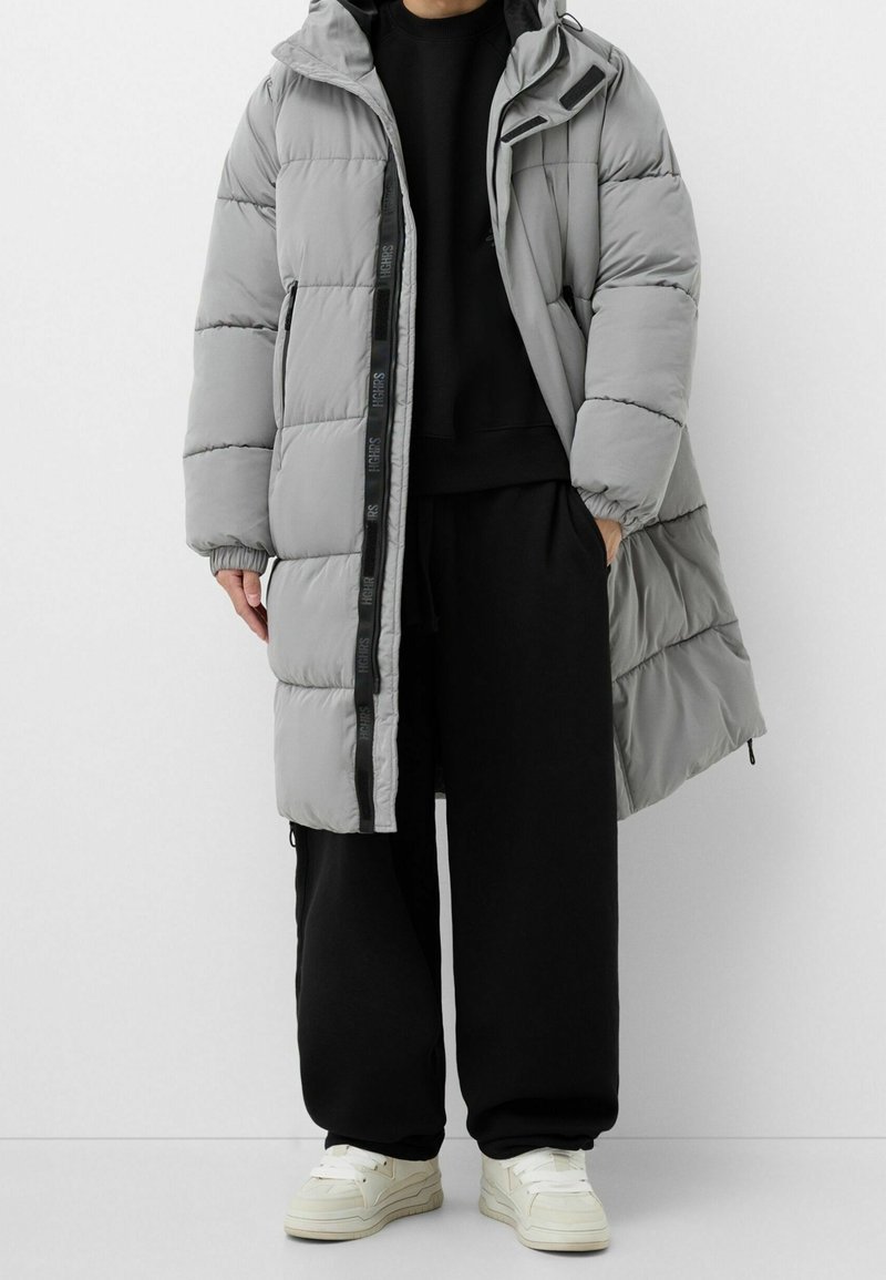 Šedý péřový kabát s kapucí, prošívaný design, přední zip a detaily s logem. Nosený přes černou mikinu a volné kalhoty.