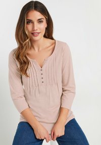 Haut à manches longues rose clair avec un col henley, présentant des plis et cinq boutons. Tissu doux et texturé avec une coupe décontractée.