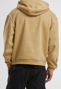 Beige mikina s kapucí, kterou nosí osoba z pohledu zezadu, v kombinaci s černými cargo kalhotami s velkými kapsami.