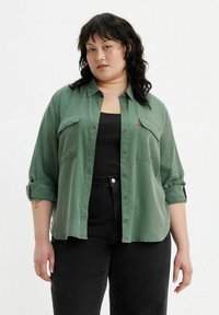 Camisa verde de botón con dos bolsillos en el pecho, mangas largas arremangadas hasta los codos y una textura suave. Combinada con una camiseta sin mangas negra y jeans.