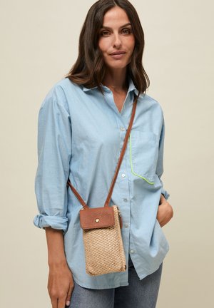 Naine, kes kannab helerohelist särki ja teksaseid, väikese kootud ristrihmaga kotiga, millel on helepruun nahast klapp ja rihm, seisab neutraalse tausta ees.