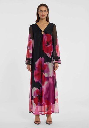 Femme portant une longue robe noire avec de grandes imprimés floraux rouges et roses, des manches longues transparentes et une découpe circulaire au niveau du décolleté.