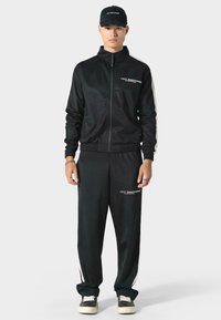 Czarny dres wykonany z gładkiego materiału, składający się z kurtki na zamek i spodni z elastycznym pasem. Białe paski po bokach i nadrukowane logo.