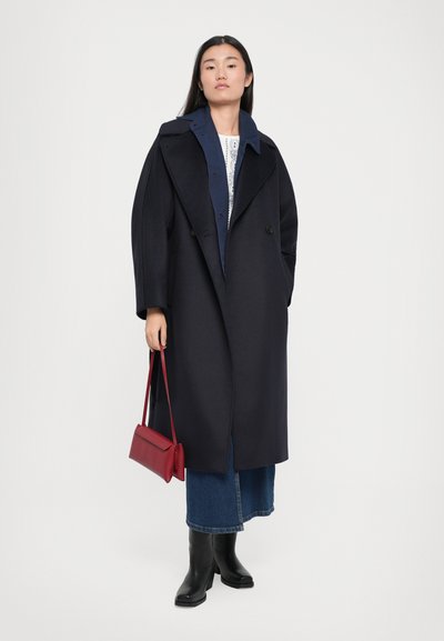 Navy oversized coat with wide lapels, two buttons, side pockets, paired with a blue top, denim skirt, and black knee-high boots, holding a red bag.