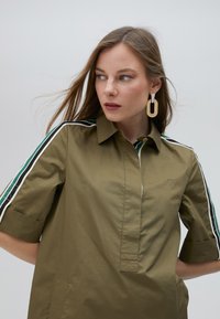 Olive green short-sleeve shirt with a collar, featuring contrasting black, white, and green stripes along the sleeves and a front placket design.