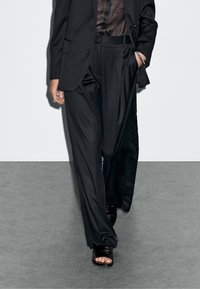 Czarny garsonka z szerokimi spodniami i przezroczystą brązową bluzką. Gładki materiał o eleganckim wykończeniu. Czarne sandały na obcasie.