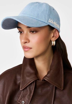 Młoda kobieta nosząca jasnoniebieską czapkę bejsbolówkę, złote kolczyki-koła oraz ciemnobrązową skórzaną kurtkę, patrzy lekko w lewo.