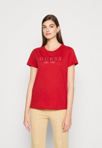 Raudona medvilninė marškinė su apvaliu iškirpimu ir trumpomis rankovėmis. Priekyje yra sidabrinių nagų logotipas "GUESS EST. 1981".