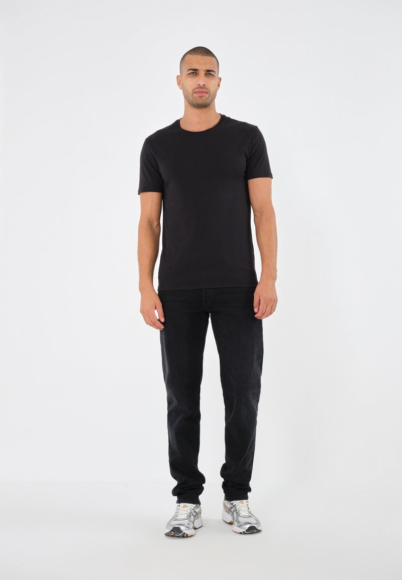 Schwarzes Kurzarm-T-Shirt, körpernah geschnitten, kombiniert mit schwarzen Jeans und gemusterten Sneakers. Minimalistisches Design auf einem schlichten weißen Hintergrund.