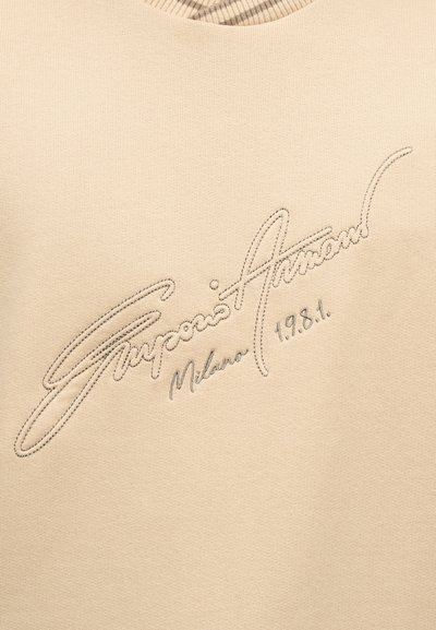 Μπεζ ύφασμα με κεντημένο κείμενο με καλλιγραφική γραφή "Giorgio Armani Milano 1981" σε νήμα ίδιου χρώματος πάνω σε υφή φόντου.