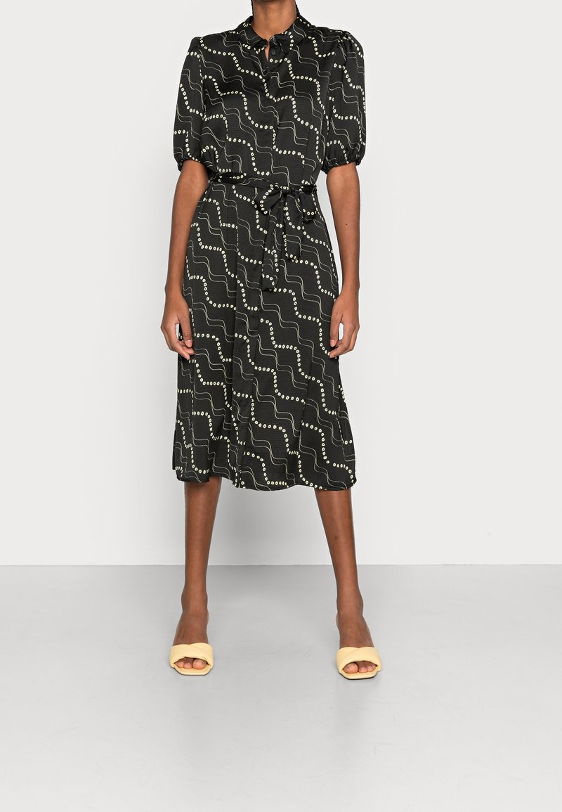 Femme portant une robe midi noire avec des motifs blancs ondulés et à pois, des manches courtes bouffantes et des sandales jaunes ouvertes aux orteils.