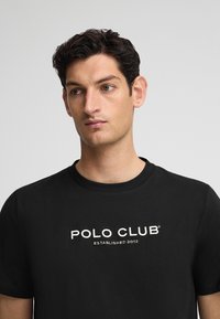 Svart bomulls t-shirt med texten "POLO CLUB" och "ETABLERAD 2012" i vit brodyr, rund halsringning, korta ärmar.