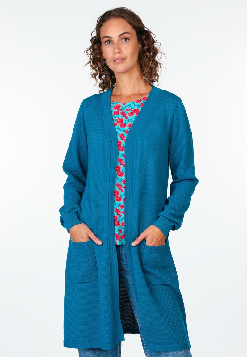 Türkiser langer Cardigan aus strukturiertem Stoff, mit offenem Front, zwei vorderen Taschen und gerippten Bündchen, über einem floral gemusterten Oberteil getragen.