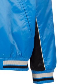 Détail de la manche et de la poche d'une veste bleue brillante avec des accents de rayures noires et beiges sur le poignet et la ceinture côtelés.