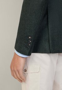 Mörkgrön texturerad kavaj med slag, fyra svarta knappar på ärmen, och en ljusblå randig skjorta synlig under ärmen.