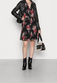 Schwarzes Blumenmuster-Kleid mit roten Blumen, kombiniert mit einer schwarzen Lederjacke und Knöchelstiefeln. Accessoires umfassen eine strukturierte Handtasche.
