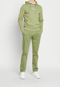 Felpa verde con tasca frontale e cordini, abbinata a pantaloni verdi coordinati. Sneakers bianche completano l'outfit casual.